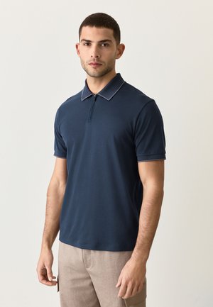 Młody mężczyzna z krótkimi ciemnymi włosami, ubrany w granatową koszulkę polo z krótkim rękawem i beżowe spodnie, stojący na tle jasnego jednolitego tła.