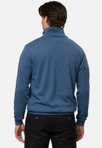 Vyras, atsuktas nugarą, dėvi mėlyną golfą ir juodas kelnes, stovintis priešais paprastą foną.