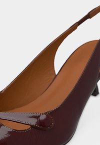 Escarpin à bride arrière en cuir verni brun foncé avec bout pointu, bride décorative et détail à bouton rond, doublure intérieure beige.