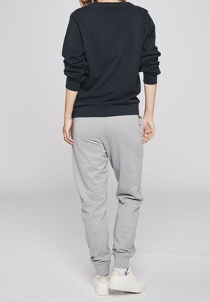 Osoba w czarnej bluzce z długim rękawem, szarych spodniach dresowych i białych trampkach, stojąca z rozluźnionymi rękami, tyłem do widza na jednolitym tle.