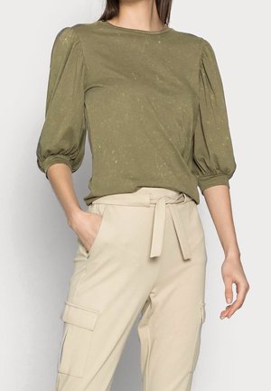 Femme portant un haut vert olive à manches bouffantes et un pantalon beige taille haute avec ceinture nouée et poches cargo latérales, debout avec une main dans la poche.