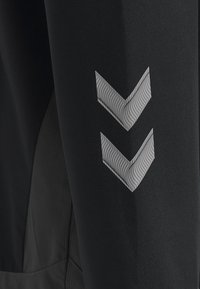 Un pantalon de sport noir présente un tissu texturé avec une finition lisse. Deux motifs en chevron de couleur gris réfléchissant sont positionnés sur le côté.