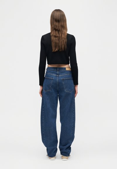 Persona con cabello largo y castaño, vestida con un top negro de manga larga corto, jeans azules holgados y zapatillas beige, de pie mirando hacia atrás sobre fondo blanco.