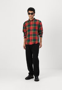 Camicia a quadri rossa e verde con bottoni, maniche lunghe, abbinata a pantaloni neri e scarpe nere, indossata con occhiali da sole scuri.