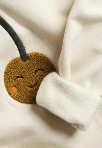 Vit tyg med en rund, brun körsbärsbroderi med ett leende ansikte, svart stjälk och mjuk textur, delvis täckt av en vikt ärm.