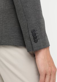 Manica di un blazer grigio con quattro bottoni neri, indossato sopra pantaloni beige chiaro, con il braccio inferiore e la mano rilassati lungo il fianco.