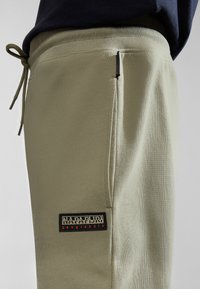 Light olive green sweatpants made of soft fabric, featuring a black patch with the brand name and logo on the side.