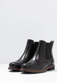 Stivali ankle in pelle nera con dettagli brogue, pannelli laterali elastici e suola marrone. Rifinitura liscia con punta rotonda e tacco basso.