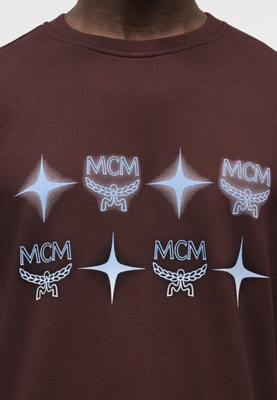 Καφέ φούτερ με επαναλαμβανόμενα λογότυπα "MCM" σε ανοιχτό μπλε και σχέδια ακτινωτής λάμψης σε στυλ μεταξοτυπίας. Κλασικός σχεδιασμός με στρογγυλή λαιμόκοψη.