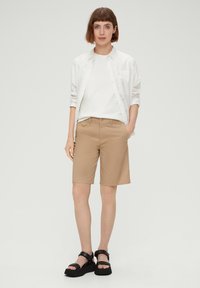 Beige shorts met een getailleerde pasvorm, gecombineerd met een wit T-shirt en een licht overhemd. Zwarte platformsandalen maken de look compleet.