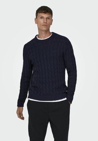 Marineblauwe kabelgebreide trui met een ronde hals en geribde manchetten en onderkant. Gedragen over een wit shirt. Zachte textuur en een ontspannen pasvorm.