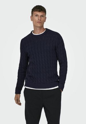 Navy cable-knit sweater with a crew neckline and ribbed cuffs and hem. Worn over a white shirt. Soft texture and relaxed fit.
