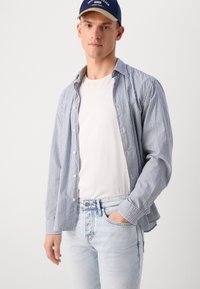 Chemise boutonnée bleu et blanc à rayures portée par-dessus un t-shirt blanc, associée à un jean bleu clair. La chemise présente une coupe décontractée et des poignets.