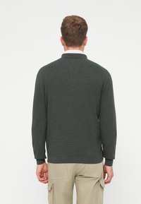 Pull vert foncé à manches longues avec col, en tissu texturé, avec des poignets et un ourlet côtelés. Un pantalon de teinte neutre est visible.