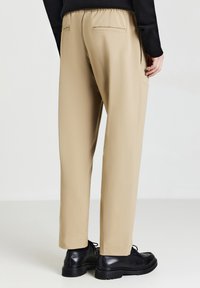 Beige Hose mit lockerem Schnitt, elastischem Bund und zwei Gesäßtaschen. Tragen mit schwarzen Schnürschuhen vor einem neutralen Hintergrund.