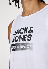 Camiseta sin mangas blanca con el texto negro en negrita "JACK & JONES" y "PERFORMANCE", con una suave textura de algodón y diseño de cuello redondo.