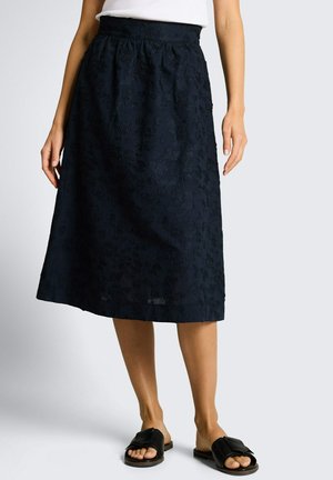 Femme portant une jupe midi noire à fleurs avec taille haute, haut blanc et sandales noires à bout ouvert, debout devant un fond clair uni.