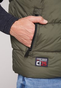 Gilet imbottito verde oliva con una tasca con zip e un'etichetta in tessuto con il marchio "CAMP DAVID". Materiale texturizzato e design aderente.