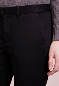 Pantalones de mezcla de algodón negros con una textura suave, con corte recto, bolsillos laterales y una cinturilla plana. Suéter de canalé gris parcialmente visible.
