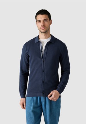 Homme portant un cardigan bleu marine boutonné sur une chemise blanche et un pantalon bleu ample, debout devant un fond gris clair uni.