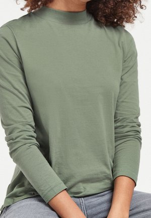 Personne portant un haut vert clair à manches longues avec col montant, assise les mains reposant sur les genoux, cheveux bouclés visibles.