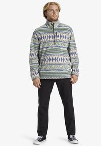 Fleece-pullover med halvlukning, der har et geometrisk mønster i blå og grøn på en cremet baggrund. Kombineret med sorte bukser og sneakers.