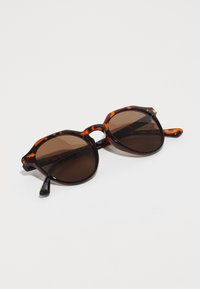 Óculos de sol tartaruga com lentes castanhas. Design de armação angular com bordas proeminentes. As hastes apresentam detalhes da marca em preto.