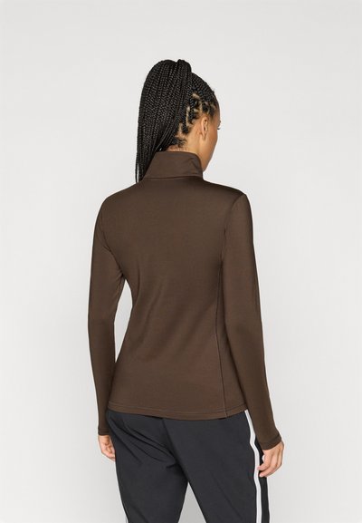 Καφέ μπλούζα μακρυμάνικη με ψηλό λαιμό, λεία υφή, πλαϊνές ραφές και εφαρμοστή σχεδίαση, συνδυασμένη με σκούρες παντελόνες που έχουν λευκές λεπτομέρειες.