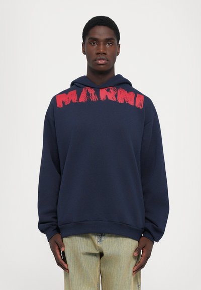 Giovane uomo che indossa un felpa blu navy con un grande testo rosso sul petto, in piedi di fronte a uno sfondo bianco semplice.