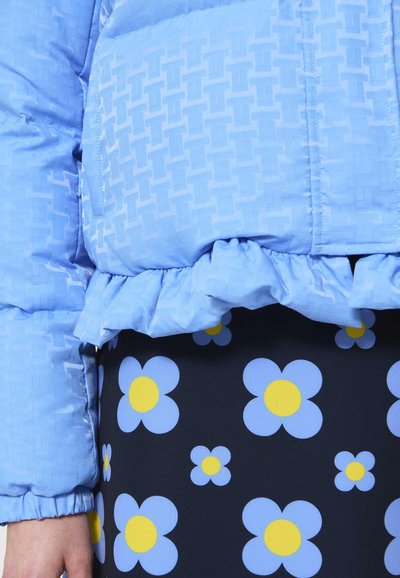 Geacă puffer de un albastru deschis, cu un model texturat și un tiv ondulat, asortată cu o fustă neagră împodobită cu imprimeuri florale albastre și galbene.