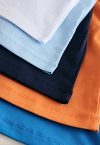 Neli puuvillast T-särki hunnikus: valge, helesinine, öösinine ja oranž. Igal särgil on ümar kaelus ning nähtav õmblusääre detailid.