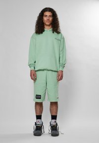 Šviesiai žalias džemperis su gobtuvu ir priekinėmis kišenėmis, derinamas su atitinkamomis šortais, turinčiais juodą logotipo užsegimą, vilkimas su juodais ir pilkais sportbačiais.