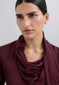 Blusa burdeos con escote drapeado. Tela suave, textura blanda y un corte holgado, con detalles doblados alrededor del cuello.