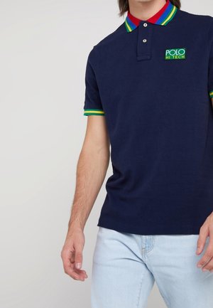 Mann trägt ein marineblaues Poloshirt mit grün-gelb-rotem gestreiftem Kragen und Ärmelbündchen, kombiniert mit hellblauer Jeans.