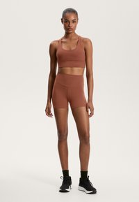 Rostfärgad sport-bh och högmidjade shorts i en slät tyg. Modellen har svarta träningsskor med mörkgröna strumpor.