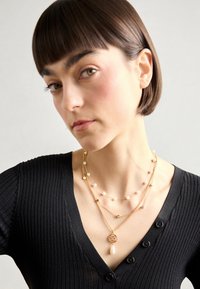 Gold-tone layered necklaces featuring delicate pearls and a circular pendant with a drop accent, worn over a black ribbed top.
