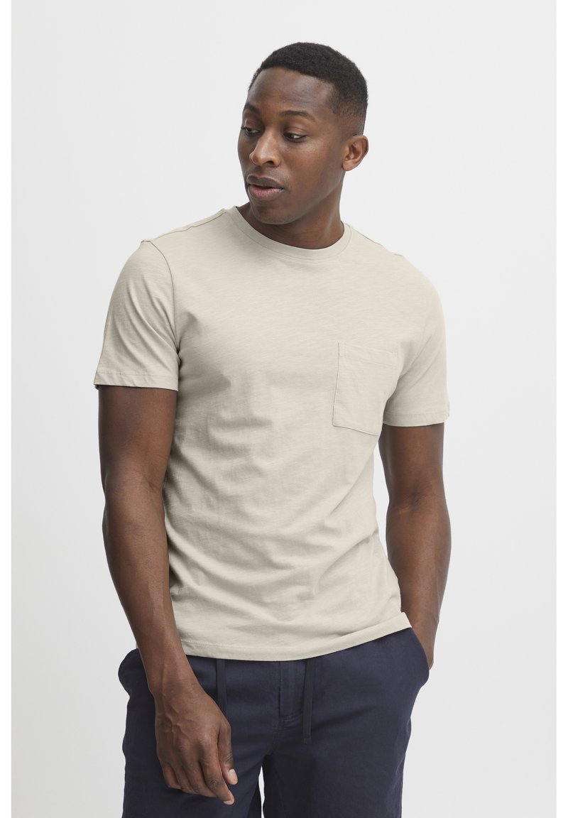 Tricou scurt cu mânecă scurtă de culoare bej deschis, realizat dintr-un material moale, având guler rotund și un mic buzunar pe piept. Modelul stă cu o expresie neutră.