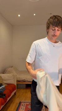 Νεαρός άνδρας με μουστάκι που διπλώνει ένα λευκό πανί σε ένα λιτά επιπλωμένο δωμάτιο με έναν καναπέ και ένα κρεβάτι.