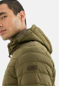 Olivgrüne Steppjacke mit quiltetem Muster, Kapuze und Knebelverschlüssen; verfügt über ein Logo-Patch auf der linken Schulter.