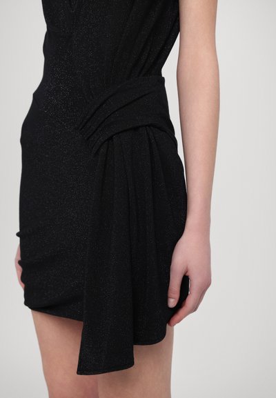 Robe noire avec une texture chatoyante, drapée de manière asymétrique et dotée d'un design noué à la taille. Un gros plan montre une main reposant sur le vêtement.