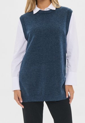 Femme portant un gilet bleu sans manches par-dessus une chemise blanche à manches longues et un pantalon noir, debout devant un fond uni.