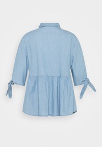 Jasnoniebieska koszula z chambray z rękawami o długości trzech ćwiartek, związywaną dekoracją przy mankietach, peplum w talii oraz klasycznym kołnierzykiem. Widok z tyłu.