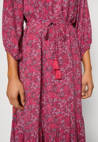 Gros plan d'une personne portant une robe florale rouge à manches longues, avec une taille cintrée et deux glands.