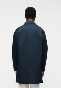 Άνδρας με κοντά, σγουρά μαλλιά που φοράει ένα σκούρο μπλε μπουφάν μεσαίου μήκους, όρθιος με την πλάτη προς τα εμπρός σε λευκό φόντο.