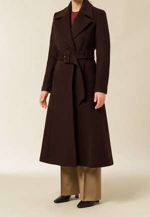 Femme portant un long manteau marron foncé ceinturé sur un pantalon beige à jambes larges et des chaussures noires à bouts pointus sur un fond neutre.
