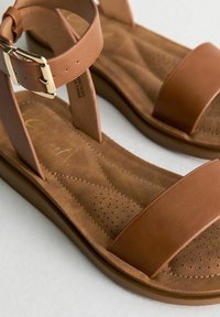 Sandales en cuir marron avec une large sangle et une boucle à la cheville, présentant une semelle intérieure texturée et des détails perforés sur la semelle.