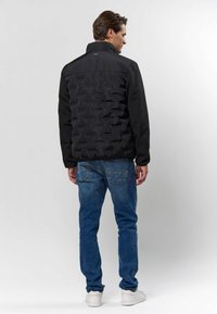 Mann som står med ryggen til, iført svart quiltet jakke, blå jeans og hvite joggesko, mot en ensfarget lys bakgrunn.