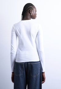 Γυναίκα με πλεγμένα μαλλιά, φοράει λευκό πουκάμισο με μακριά μανίκια και μπλε τζιν, αντιμετωπίζει την κάμερα με την πλάτη σε απλό λευκό φόντο.