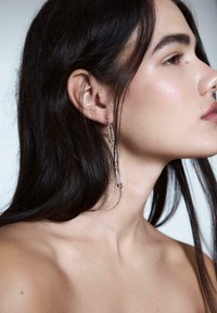 Profilo di una persona con lunghi capelli scuri che indossa un orecchino d'argento pendente fatto di spille da balia collegate, su uno sfondo semplice.