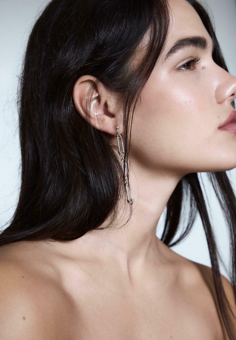 Profilo di una persona con lunghi capelli scuri che indossa un orecchino d'argento pendente fatto di spille da balia collegate, su uno sfondo semplice.